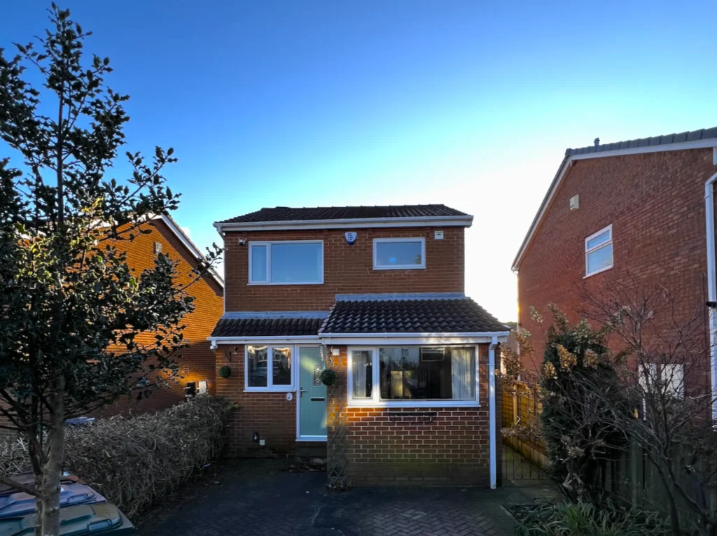 A two-story brick house with a tiled roof, surrounded by trees and adjacent to two other houses, against a clear blue sky.